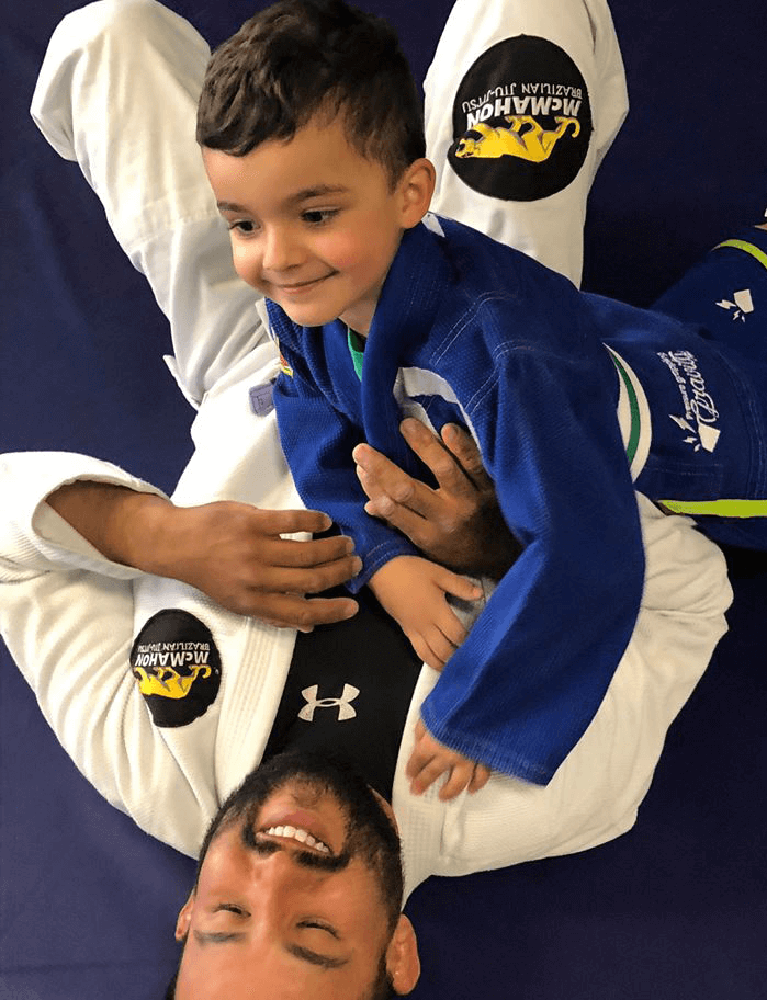 child in blue brazilian jiu-jitsu gi in side control on top of his father in white gi